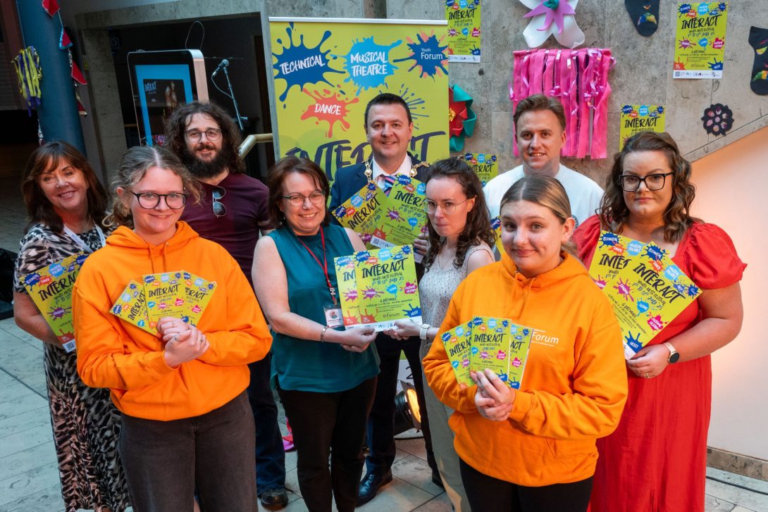 Group of people at the Launch of InterAct Youth Arts Festival, some holding colourful flyers for the Millennium Forum's 'InterAct Youth Arts Festival'. Two individuals in bright orange hoodies stand at the front, with others behind them smiling in casual and business attire. A vibrant poster and a decorated wall are visible in the background.