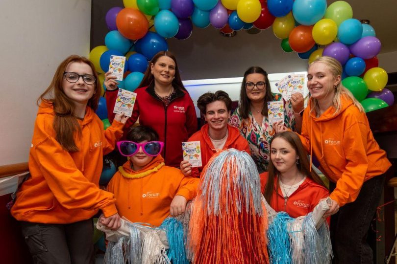 Group of young people in bright orange hoodies posing under a colourful balloon arch, holding flyers and smiling, with one person in fun costume and oversized sunglasses.