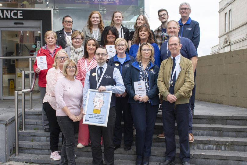 A group of staff and partners pose on the steps outside the Millennium Forum, holding certificates and promotional materials, smiling for a photo to mark a community or accessibility award.