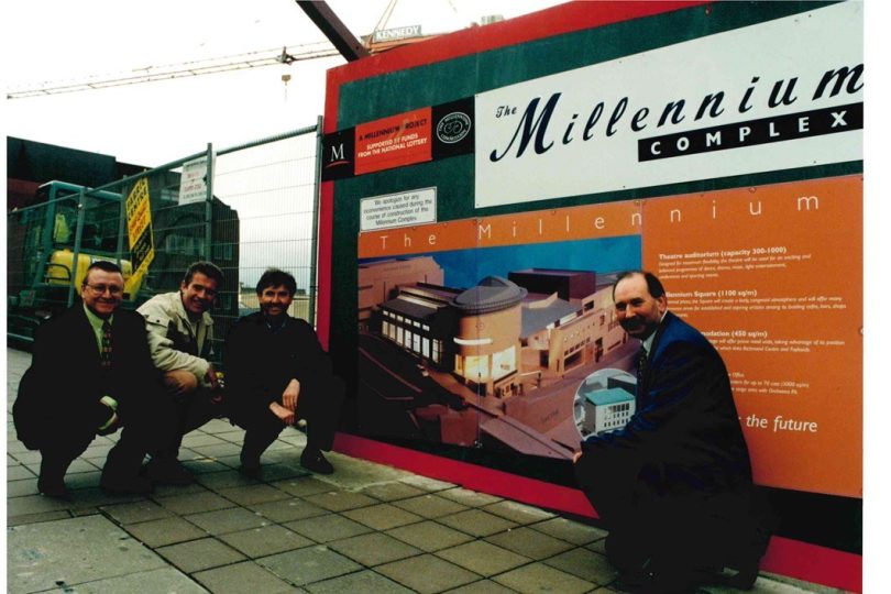 Four men crouch and smile in front of a construction hoarding featuring design plans for the Millennium Forum, with cranes and building works visible in the background.