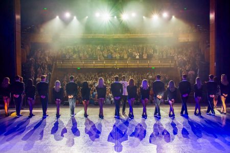 Finale scene from Lord of the Dance, showing the full cast of dancers standing in a line on stage facing a cheering audience. The performers are backlit with dramatic stage lighting, casting long shadows across the floor as they take their bows.