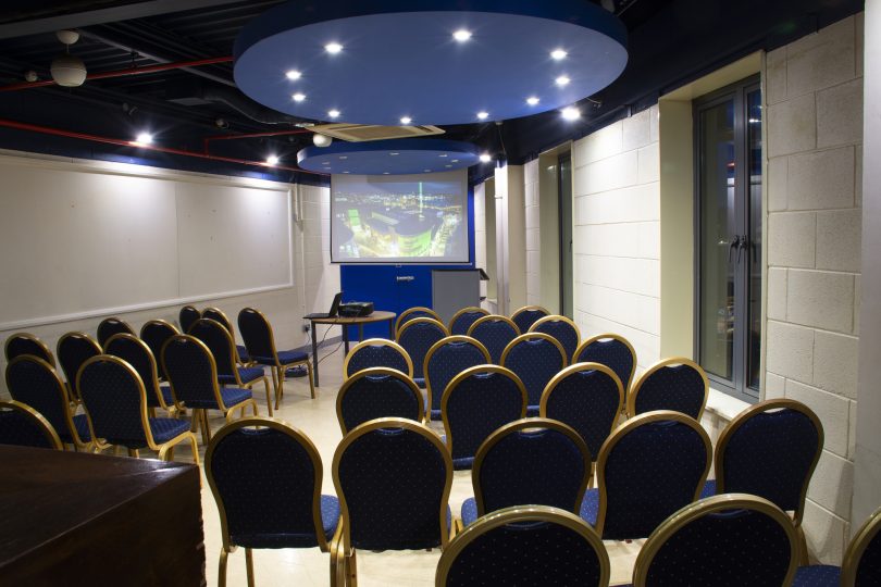 Our green room with navy and gold chairs arranged in theatre style facing a screen displaying a presentation. The room features white walls, spotlight ceiling lighting, and a blue circular ceiling feature above the front podium.