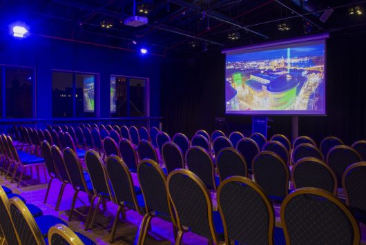 Rows of blue and gold chairs set up for a presentation in a darkened room, with a large screen displaying a colourful aerial image of Derry lit up at night.