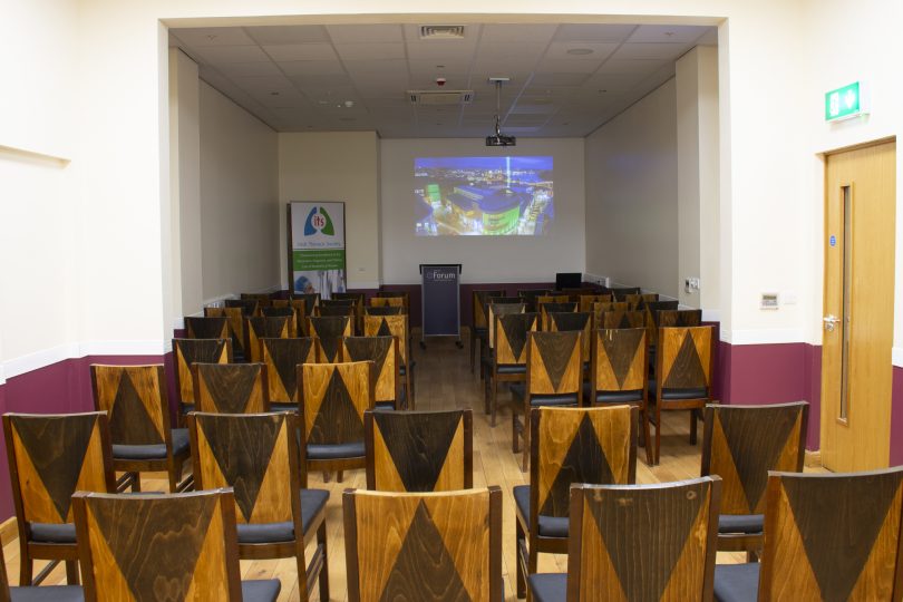 A small conference room set up with wooden chairs facing a podium and projection screen, ready for a presentation or seminar, with branding from the Irish Thoracic Society displayed on a banner.