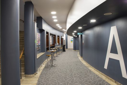 Modern theatre corridor with curved walls, grey carpet, bar tables with stools, and signage indicating ‘A’ for the auditorium door A entrance. Well-lit with ceiling spotlights and access to staircases.