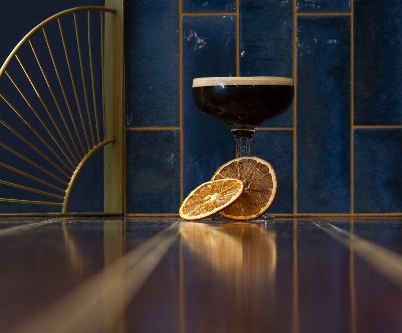 A stylish espresso martini garnished with dried orange slices, served on a glossy wooden bar with a navy and gold-tiled backdrop and art deco-style gold railing detail.