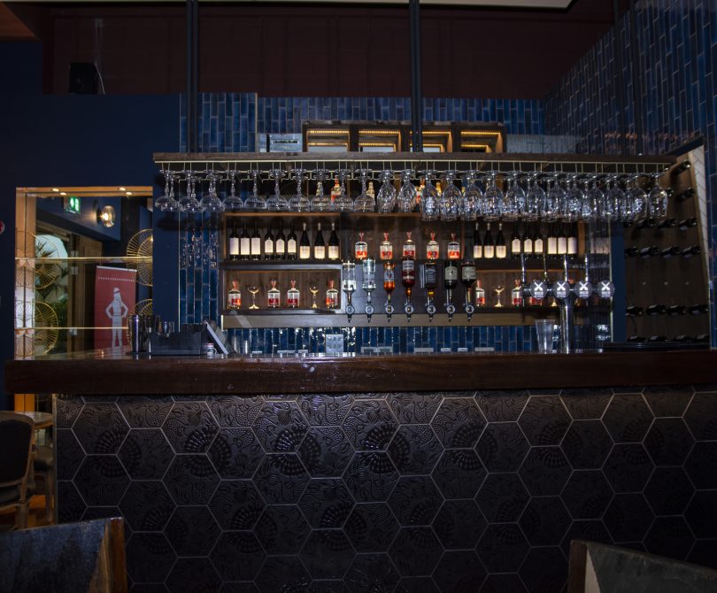 Stylish bar interior featuring a dark hexagonal-tiled front, hanging wine glasses, and a backlit display of premium spirits against a blue tiled wall.