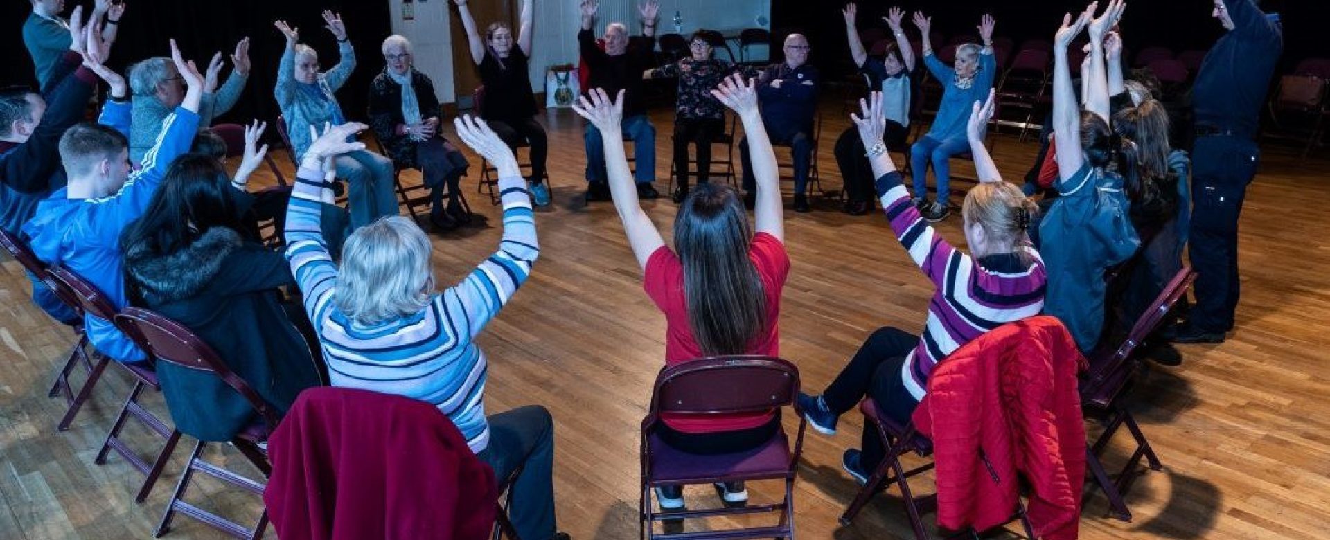 A group of people sit in a large circle on chairs in a spacious room, all raising their arms enthusiastically during a group activity session on a wooden floor.