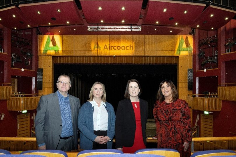 Four people stand inside the Millennium Forum auditorium with the stage in the background. The Aircoach logo is prominently displayed on the proscenium arch above the stage.