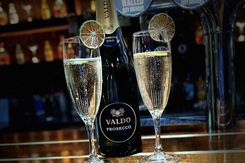 A bottle of Valdo Prosecco and two filled champagne flutes garnished with lime slices, set on a wooden bar counter with beer taps and spirits in the background.