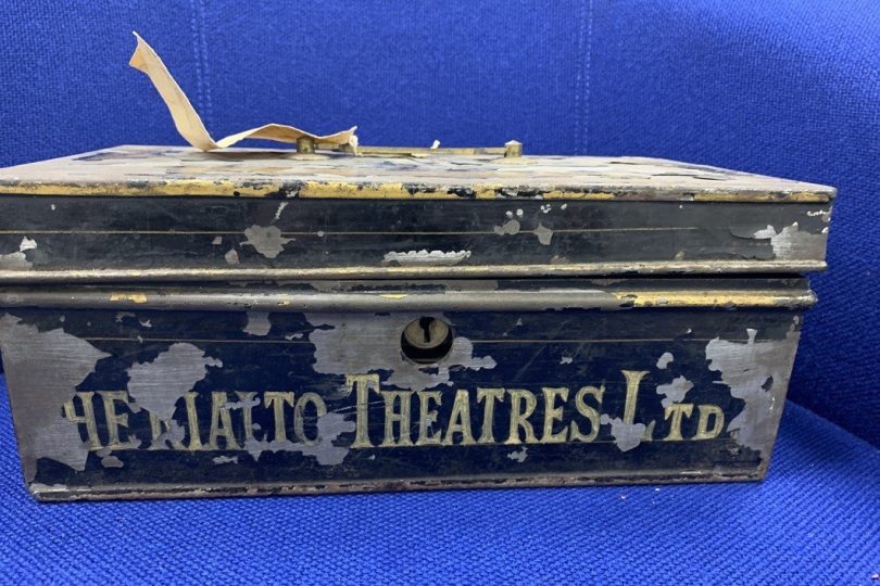 Old, weathered metal cash box with faded text reading “The Rialto Theatres Ltd”, placed against a blue fabric background.