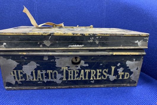 Old, weathered metal cash box with faded text reading “The Rialto Theatres Ltd”, placed against a blue fabric background.