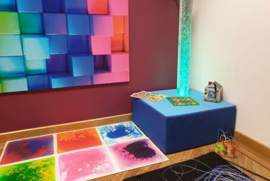 Sensory room featuring a soft blue seating block, interactive light-up bubble tube, colourful wall panels, sensory floor tiles, and tactile toys designed for neurodiverse children and young people.