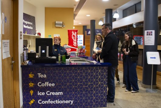 Customers are being served at the Millennium Forum’s refreshments kiosk, with signage promoting tea, coffee, ice cream, and confectionery. The counter has a geometric blue and gold design, and the area is well-lit with branded fridges and product displays in the background.