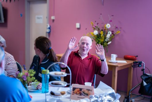 A smiling man in a red polo shirt raises both hands in a lively gesture while seated at a community tea event table, with cakes, flowers, and other attendees in the background.