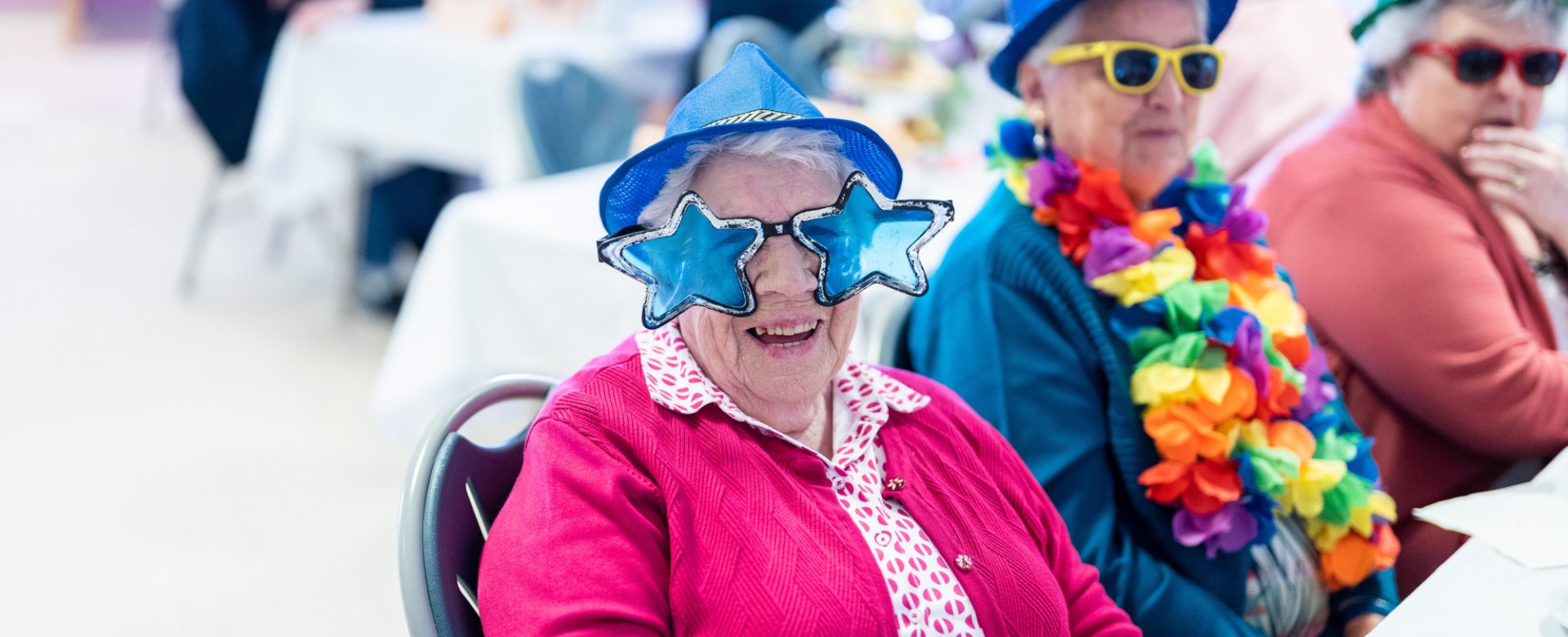 Older adults dressed in colourful party accessories, including star-shaped glasses, hats, and feather boas, smile and enjoy a community tea dance or celebration event.