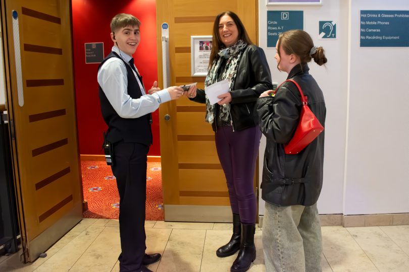 Millennium Forum front-of-house staff member smiling while scanning a guest’s ticket at the entrance to the auditorium, with two audience members preparing to enter.