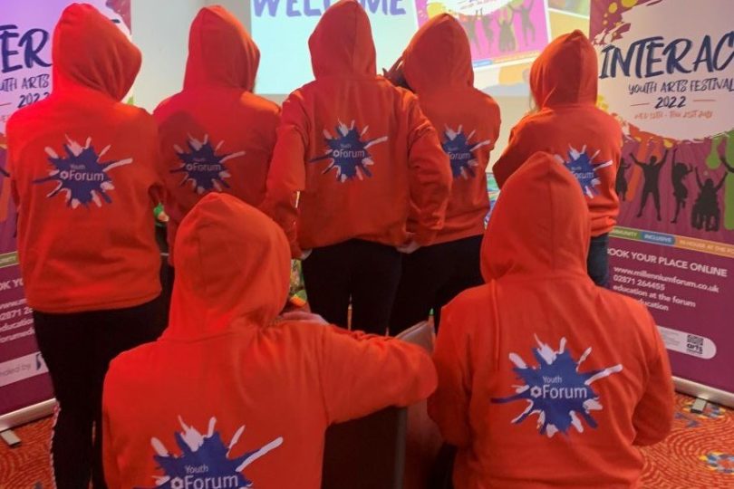 Group of young people wearing matching red hoodies with the Youth Forum logo on the back, facing a projected screen that reads “Welcome to InterAct”, with festival banners on either side.