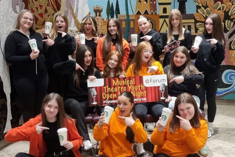 Group of smiling young people, some in orange hoodies, holding drinks and posing enthusiastically with a sign that reads “Musical Theatre Youth Open Night”, in front of a colourful mural backdrop.