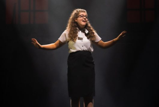 A young female performer stands centre stage under a dramatic spotlight, mid-performance, with arms raised slightly and an expressive facial gesture.