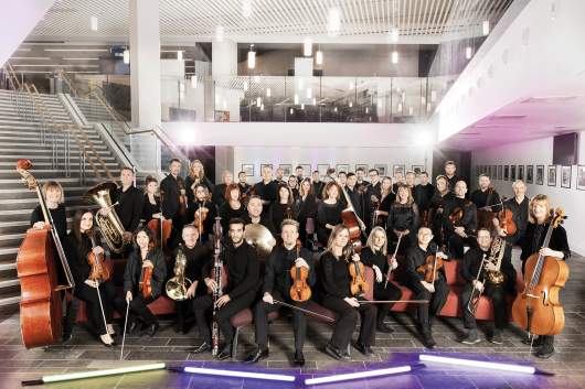 Group photo of a full orchestra posed in a modern indoor venue with sleek architecture, featuring a staircase and mezzanine in the background. The musicians are dressed in black concert attire and arranged in rows, holding their instruments including strings, woodwinds, brass, and percussion. The setting is brightly lit with a polished floor and ambient ceiling lights, giving the image a professional and elegant atmosphere, highlighting the scale and unity of the ensemble.