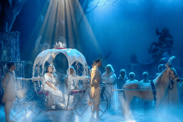 Scene from the Cinderella pantomime featuring Cinderella in a sparkling white gown seated in a magical pumpkin-shaped carriage, drawn by a white horse. She is surrounded by footmen and performers dressed in white, with soft blue lighting and stage fog creating an enchanting fairy-tale atmosphere.