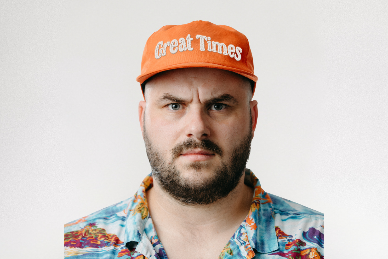 A man with a serious expression wearing an orange cap that says ‘Great Times’ and a colorful patterned shirt, standing against a plain light background