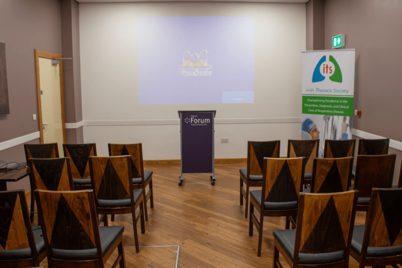 Small conference room setup at the Millennium Forum with rows of wooden chairs facing a podium and projection screen. A banner for the Irish Thoracic Society stands to the side.