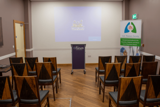 Small conference room setup at the Millennium Forum with rows of wooden chairs facing a podium and projection screen. A banner for the Irish Thoracic Society stands to the side.