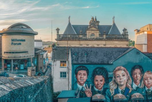 View of the Millennium Forum theatre in Derry, Northern Ireland, beside a large mural of the Derry Girls characters, with historic architecture and a blue sky in the background.