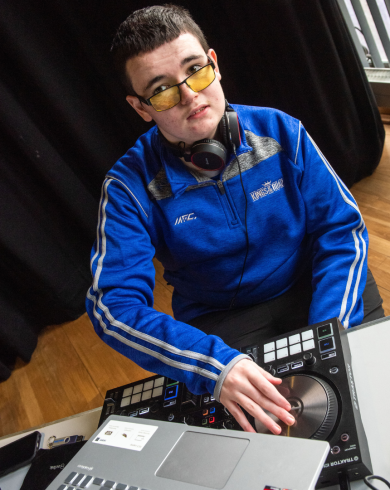 A young person in a blue tracksuit and yellow-tinted glasses using DJ equipment, wearing headphones around their neck and standing beside a laptop in a performance or workshop setting.
