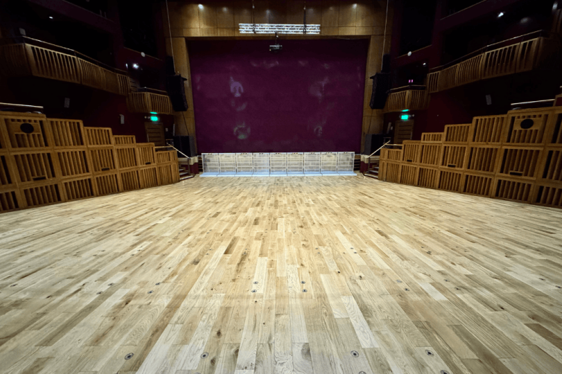 Wide-angle view of an empty auditorium at the Millennium Forum with polished wooden flooring, red walls, and wooden side balconies.