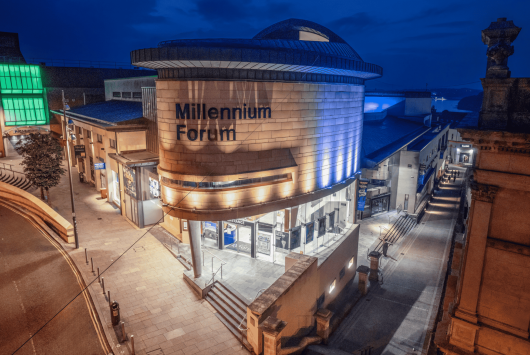 Night-time view of the Millennium Forum in Derry, with its curved stone facade illuminated by blue and white lighting, and surrounding pedestrian walkways.