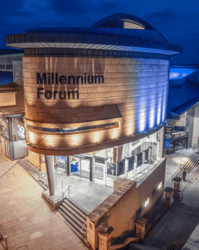 Night-time view of the Millennium Forum in Derry, with its curved stone facade illuminated by blue and white lighting, and surrounding pedestrian walkways.