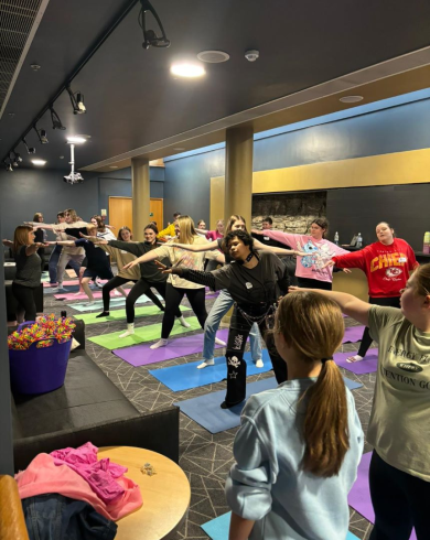A group of people participating in a yoga or fitness class indoors, stretching on colourful mats with an instructor leading the session in front.