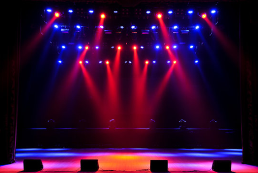 An empty stage illuminated by dramatic red and blue stage lights, with several microphones positioned at the front, ready for a live performance.