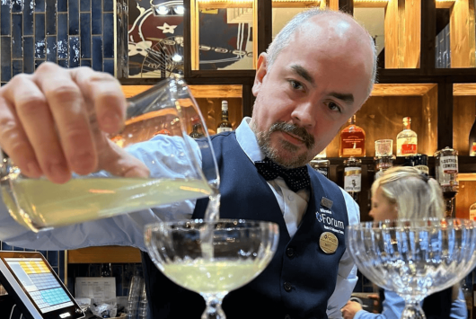 A bartender at the Millennium Forum's Speakeasy bar pours a cocktail into a glass. He is dressed in a blue waistcoat, bow tie, and name badge, with shelves of spirits and a stylish tiled wall in the background.
