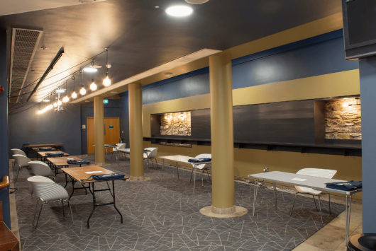 East Wall bar area at the Millennium Forum with folding tables, cushioned chairs, feature lighting, and a geometric-patterned carpet. The room has navy and gold decor with exposed stone detailing.