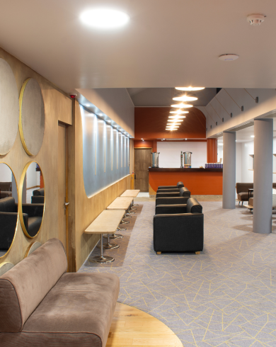 Modern theatre lounge area featuring stylish seating, circular wall mirrors, bench seating along the wall, and warm ambient lighting leading to a reception or bar area.