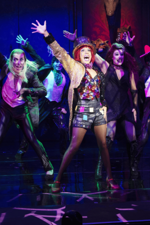 Energetic stage scene from The Rocky Horror Show featuring a vibrant central performer in a sequinned top, gold jacket, and red hat, surrounded by dancers in gothic and eccentric costumes. All performers strike dramatic poses under colourful, theatrical lighting.