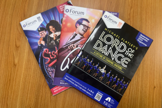 Three Millennium Forum show programmes laid out on a wooden surface, featuring Sister Act, The Elvis Spectacular, and Lord of the Dance.