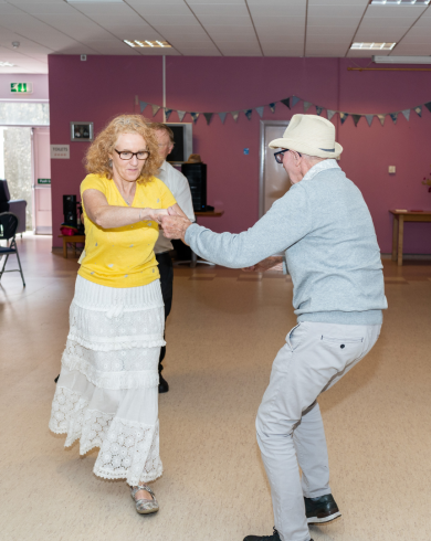 An older couple dancing together in a brightly lit community room with pink walls and bunting. The woman wears a yellow top and white skirt, and the man wears a light blue jumper, grey trousers, and a white hat.