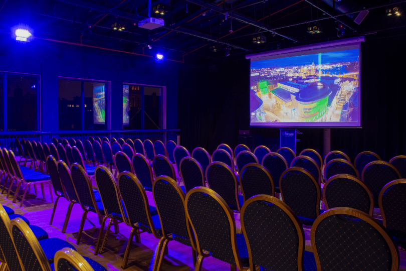 A modern presentation room with rows of blue cushioned chairs facing a projector screen displaying a vibrant cityscape, illuminated by purple ambient lighting.