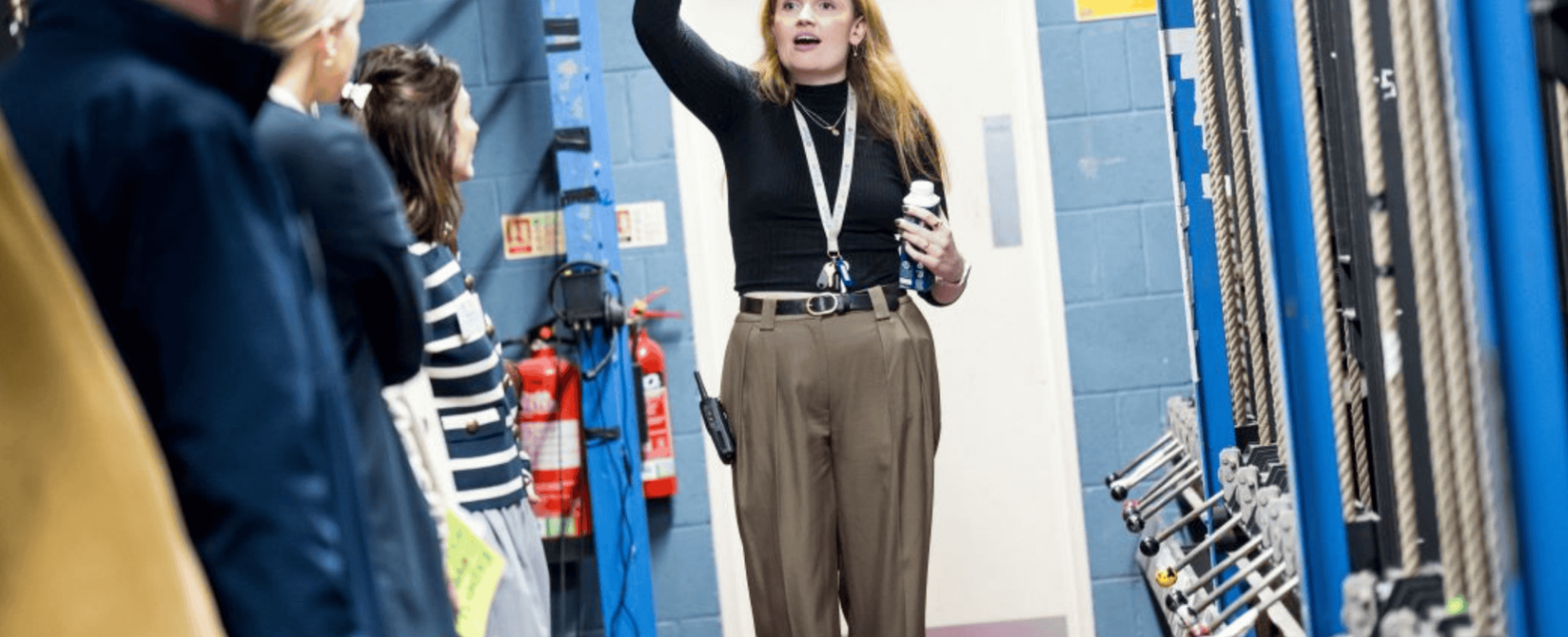 A female theatre technician leads a backstage tour, gesturing upwards while explaining the fly system to a group of visitors standing in a technical corridor with rigging ropes and fire extinguishers visible.