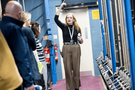 A female theatre technician leads a backstage tour, gesturing upwards while explaining the fly system to a group of visitors standing in a technical corridor with rigging ropes and fire extinguishers visible.