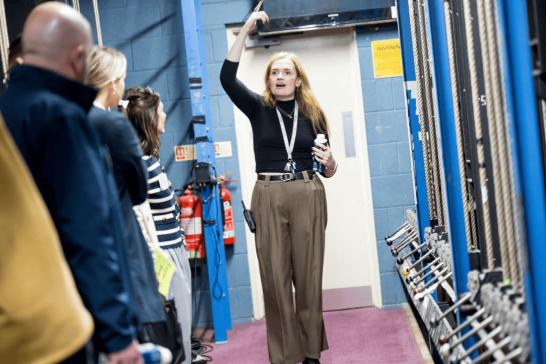 A female theatre technician leads a backstage tour, gesturing upwards while explaining the fly system to a group of visitors standing in a technical corridor with rigging ropes and fire extinguishers visible.