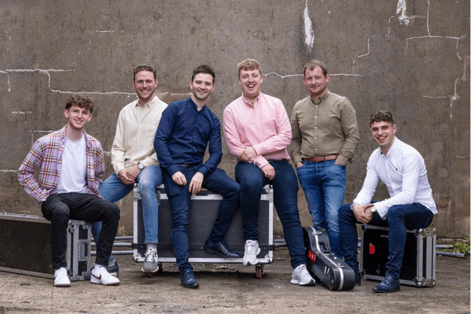 Promotional image of The Tumbling Paddies, a six-piece Irish band. The members are casually dressed in jeans, shirts, and trainers, sitting and standing in front of a rustic concrete wall. They’re posed around instrument flight cases and a guitar case, hinting at their musical setup. Their relaxed, approachable energy reflects the band’s upbeat, contemporary take on Irish folk and country music.