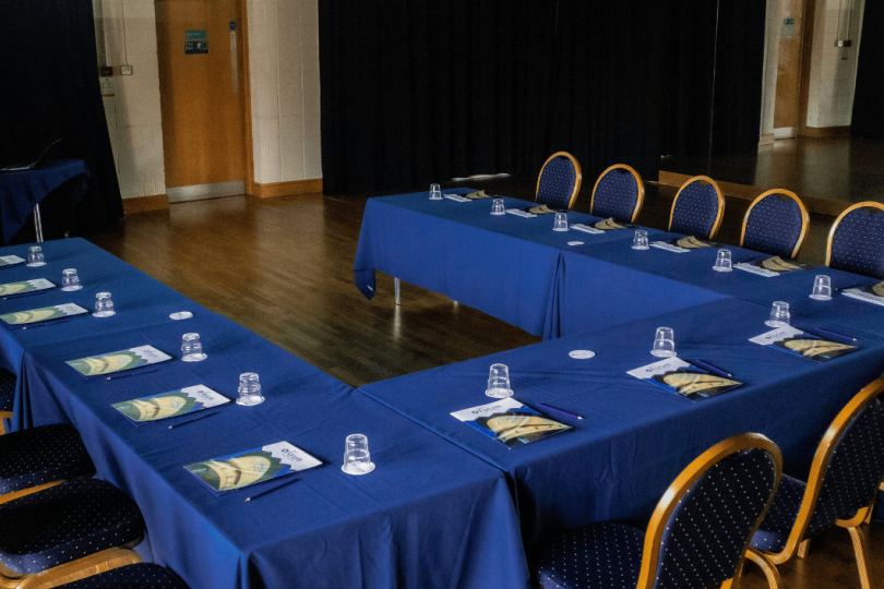 A U-shaped conference table setup with navy blue tablecloths and matching cushioned chairs, each place setting equipped with a glass, a pen, and printed materials, in a professional meeting room.