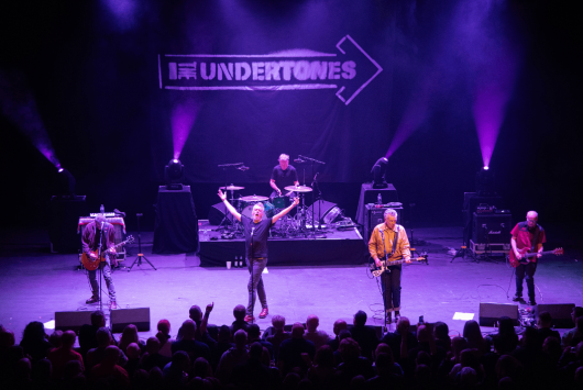 The Undertones performing live on stage at the Millennium Forum, with vibrant purple lighting and a crowd watching from the auditorium. A large banner with the band’s name hangs behind the drum kit.