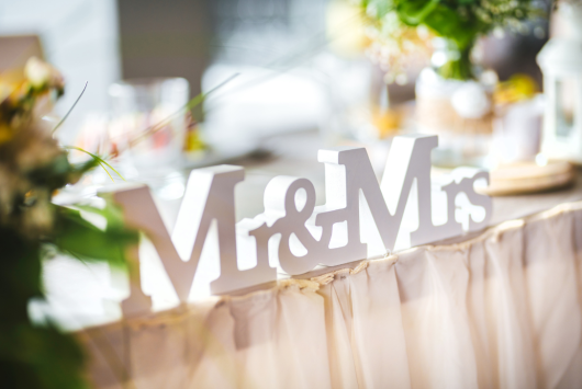 Close-up of a white “Mr & Mrs” sign on a softly decorated wedding table, with blurred floral arrangements and glassware in the background.
